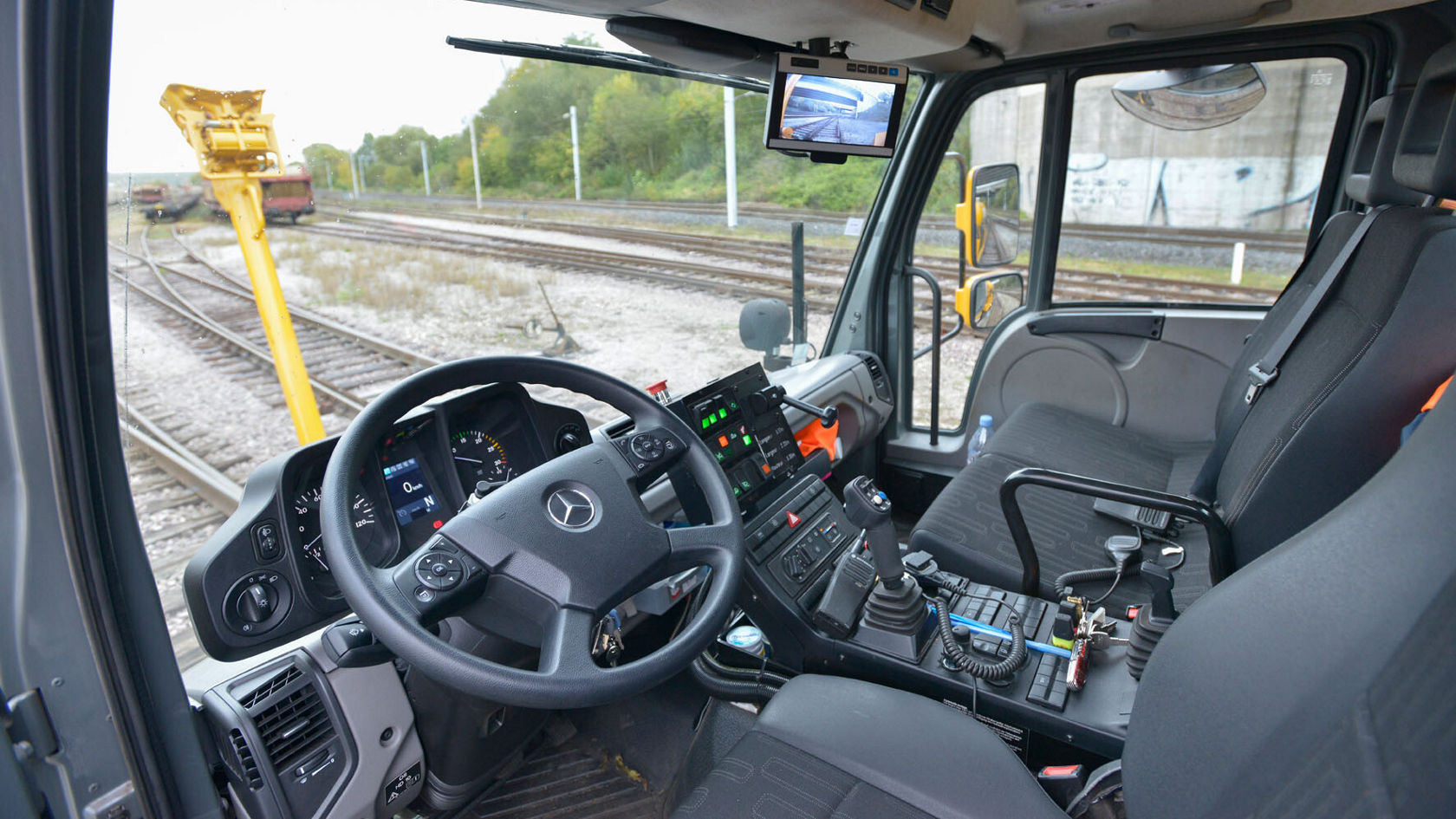 Innenansicht eines Mercedes-Benz Unimog mit einem Lenkrad, Bedienelementen und einem Bildschirm, der die Schienenlandschaft zeigt.