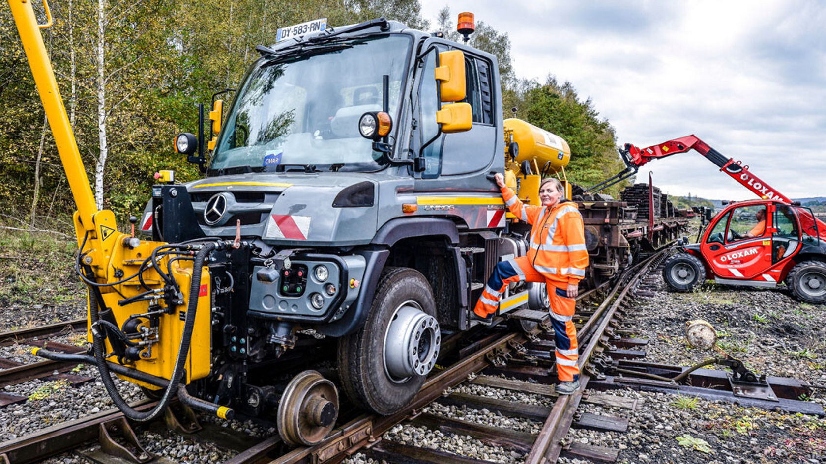 Ein Mercedes-Benz Unimog steht auf einem Gleis, während ein Arbeiter in einer orangefarbenen Sicherheitsjacke daneben steht. Im Hintergrund ist ein weiteres Fahrzeug zu sehen.