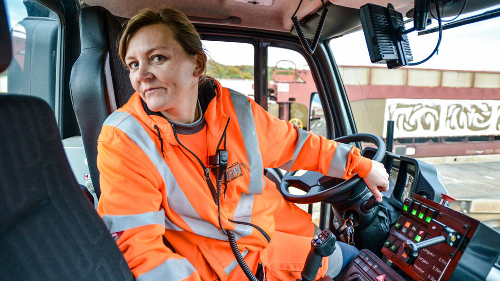 Eine Person in leuchtend orangefarbener Arbeitskleidung sitzt im Fahrersitz eines Mercedes-Benz Unimog und steuert die Bedienelemente des Fahrzeugs.