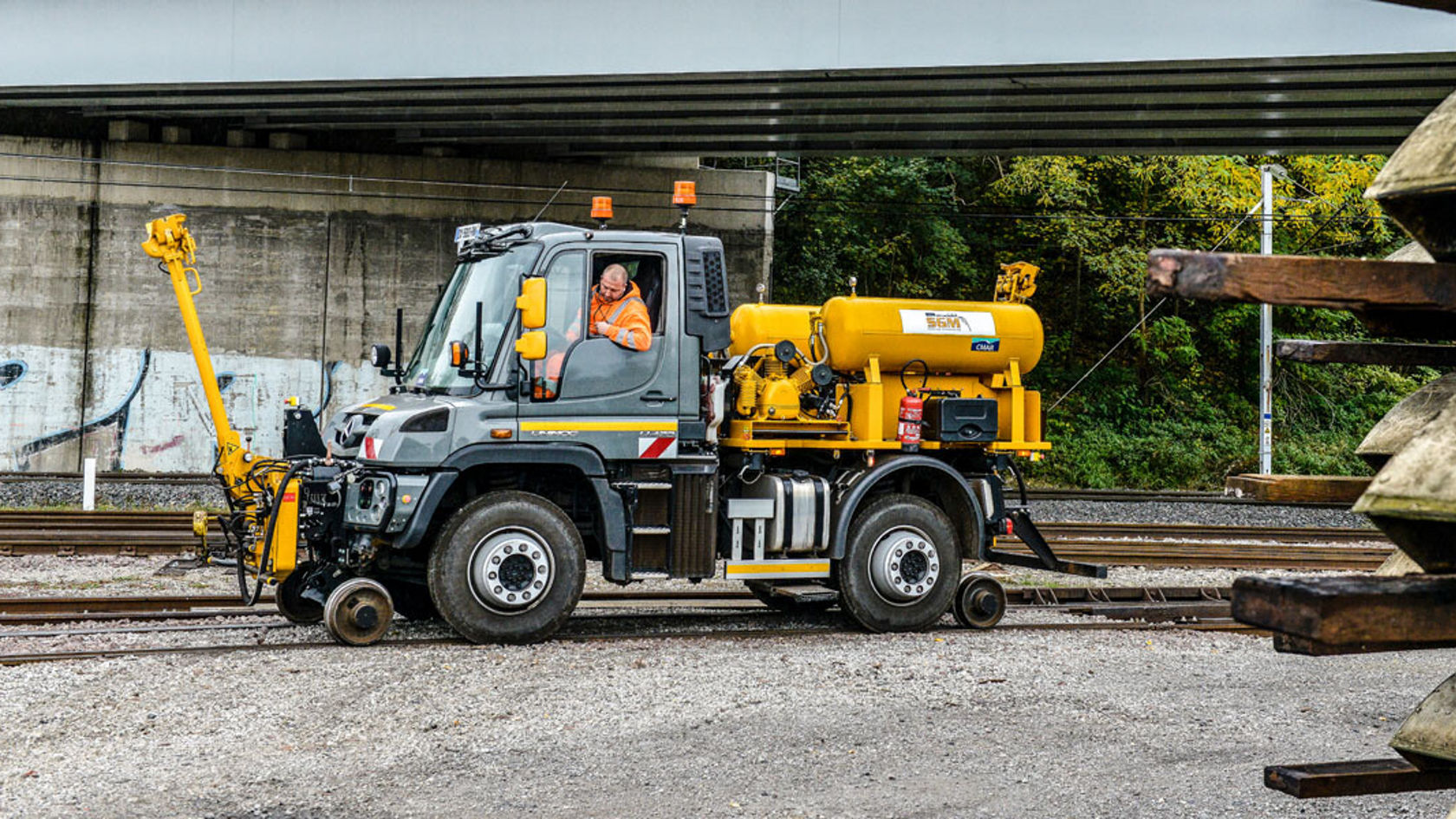 Ein Mercedes-Benz Unimog steht auf einer Bahnstrecke, ausgestattet mit einem Gelbkompressor und einem Arbeitsarm. Ein Fahrer ist im Fahrersitz zu sehen.