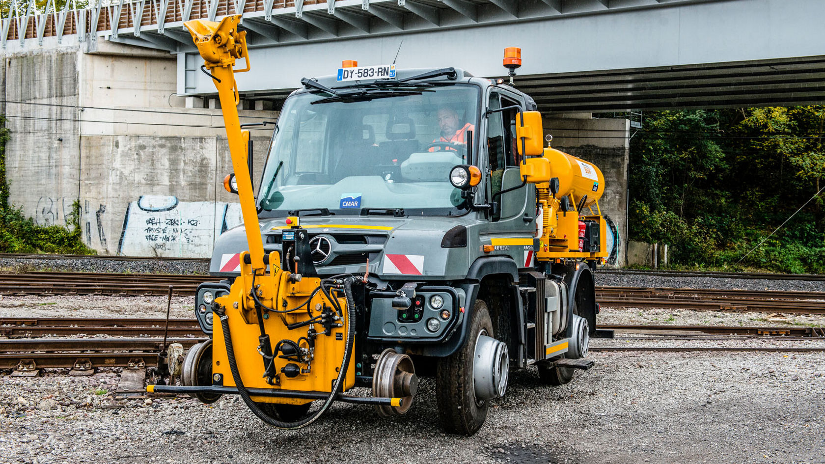 Ein gelber Mercedes-Benz Unimog mit einer Anbauvorrichtung steht auf einem Gleis zwischen Bahngleisen, bereit für Wartungsarbeiten.
