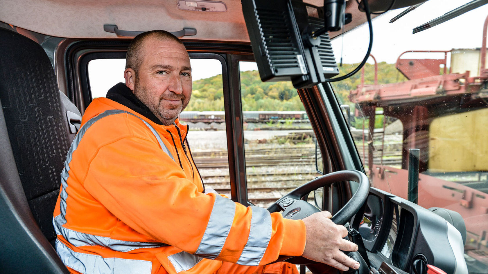 Ein Mann in einer Warnjacke sitzt am Steuer eines Unimog von Mercedes-Benz. Er lächelt und schaut zur Kamera, während er das Fahrzeug steuert.