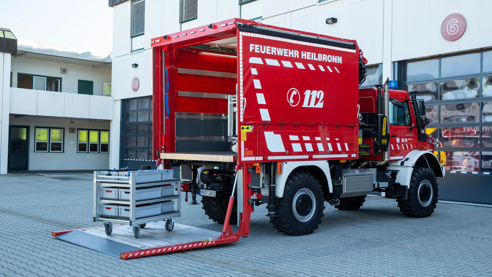 Un véhicule de pompiers rouge des sapeurs-pompiers de Heilbronn, avec la zone de chargement ouverte révélant un rayonnage coulissant. L'avant du véhicule est bien visible, avec le numéro d'appel d'urgence « 112 ».