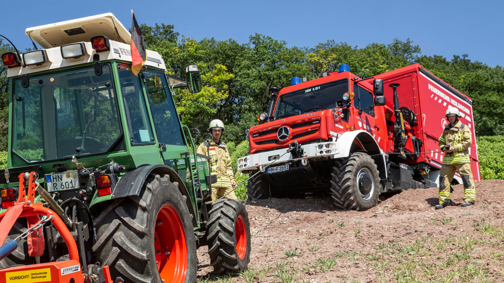 Un camion de pompiers Mercedes‑Benz se trouve dans un champ à côté d'un tracteur. Deux pompiers en uniforme se tiennent à proximité du camion.