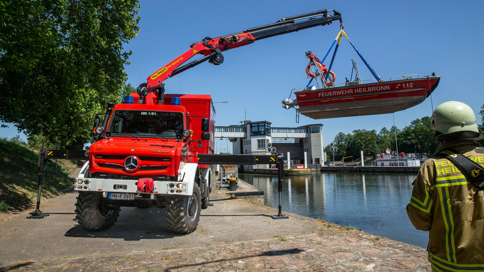 Un véhicule d'incendie rouge de Mercedes‑Benz soulève à l'aide d'une grue un bateau de sauvetage au‑dessus du canal, tandis qu'un pompier en uniforme observe.