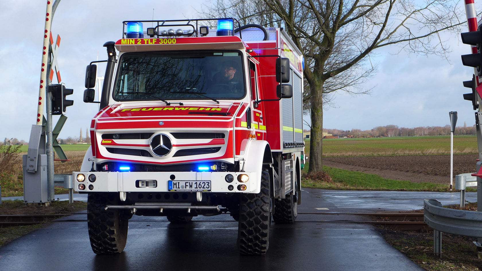 Un camion de pompiers Mercedes‑Benz rouge, gyrophare allumé, est arrêté devant une barrière de passage à niveau. Le conducteur est visible, tandis que le camion est stationné sur une route.