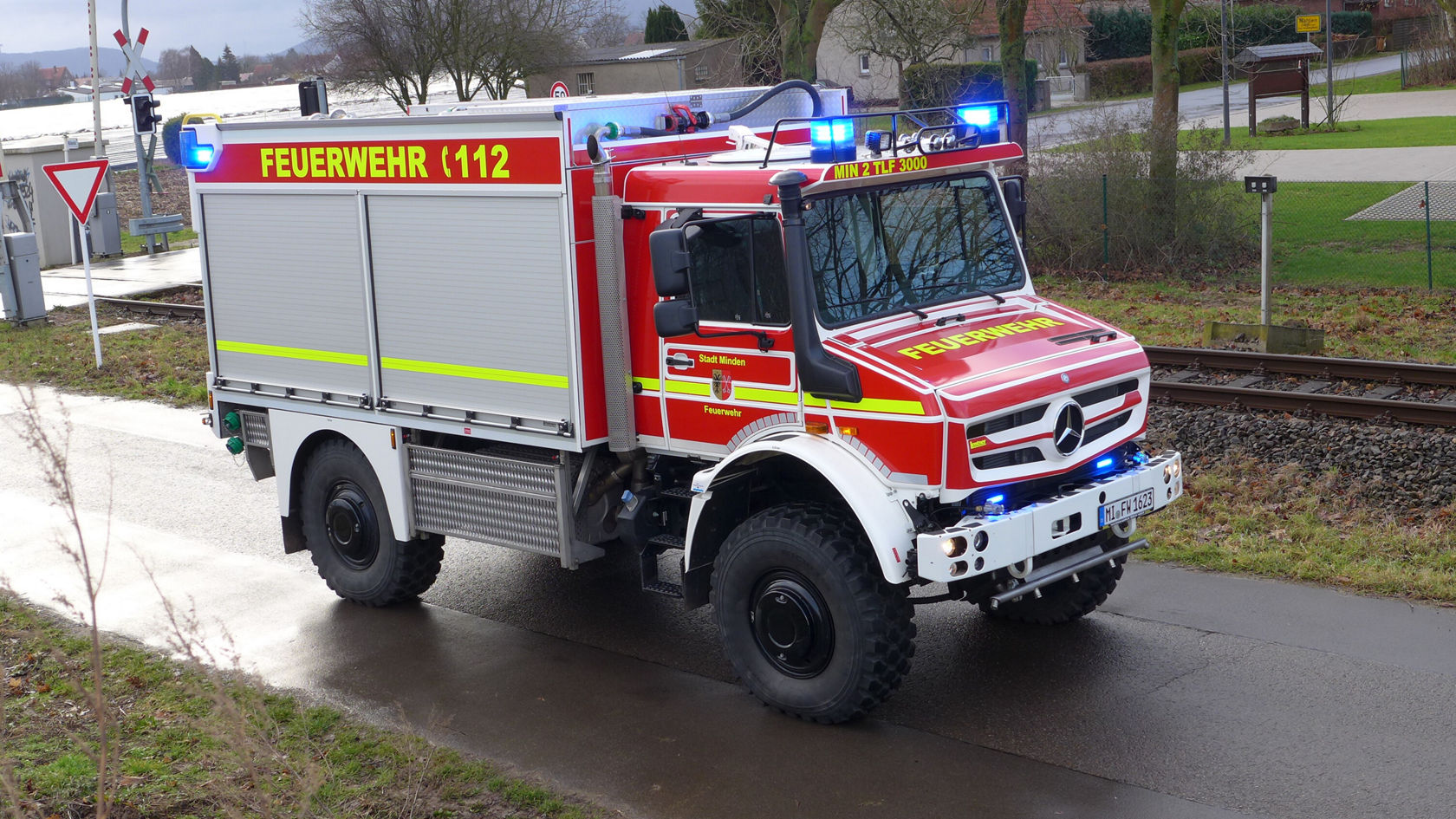 Un camion de pompiers Mercedes‑Benz rouge circule sur une route, avec les feux bleus allumés. Sur le flanc, on lit « FEUERWEHR 112 » en lettres jaunes.