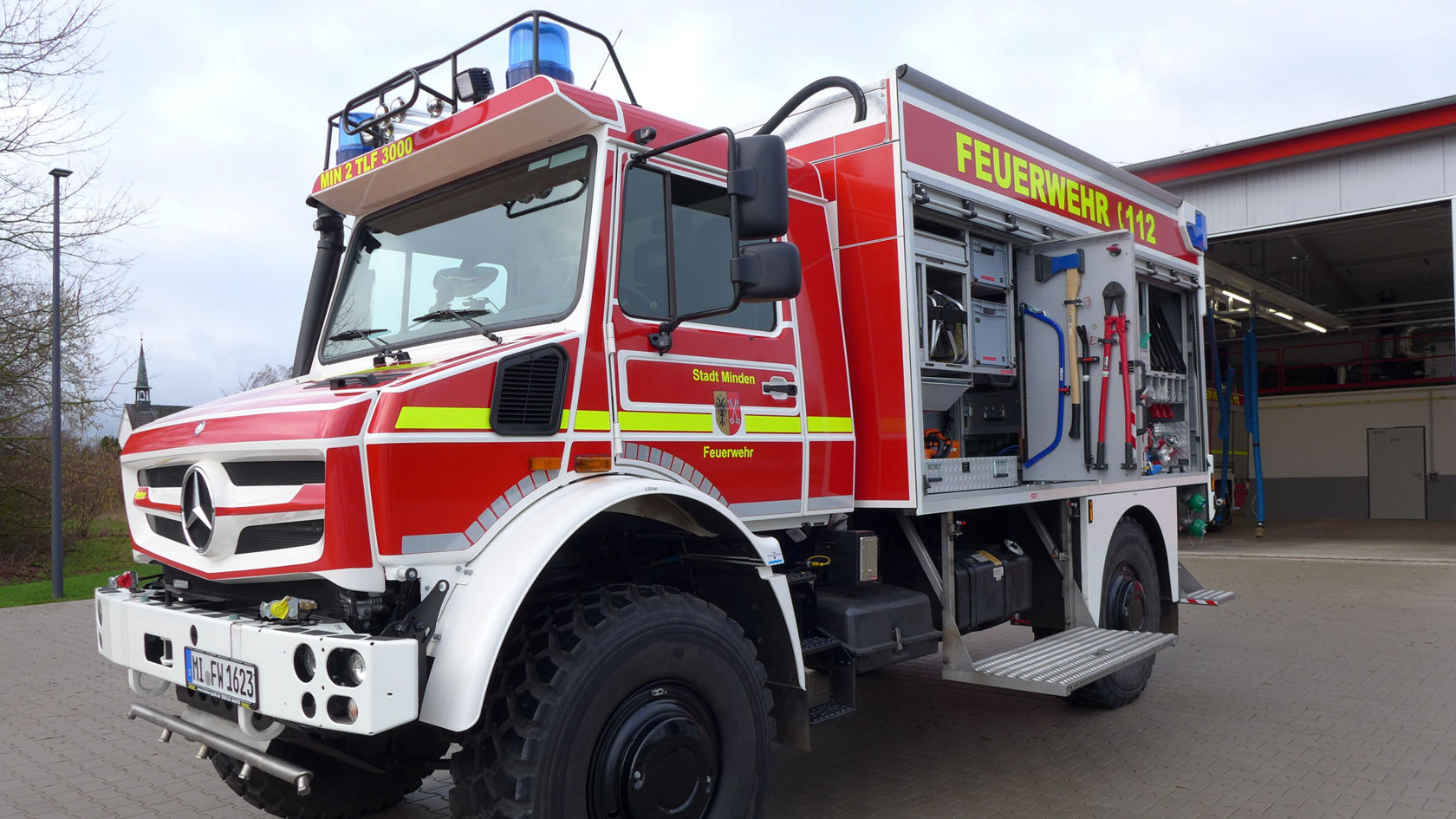 Un camion de pompiers Mercedes‑Benz, peint en rouge avec des bandes jaunes et équipé de matériel de lutte contre les incendies, est stationné devant une caserne de pompiers.