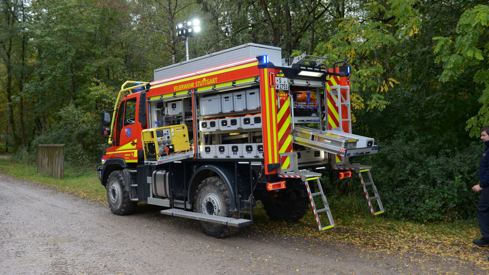 Ein Mercedes-Benz Feuerwehrfahrzeug steht an einem Waldweg. Der Fahrzeugaufbau ist mit verschiedenen Ausrüstungen und Lampen ausgestattet, bereit für den Einsatz.