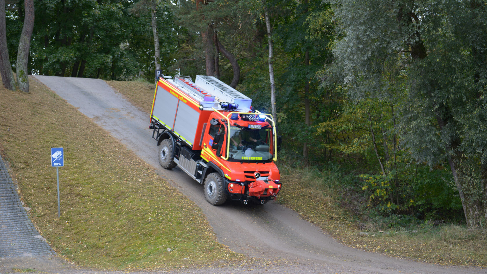 A Mercedes-Benz fire engine is driving up a gravel road, surrounded by trees and grass. It has its blue lights on and is painted in conspicuous red and yellow colours.