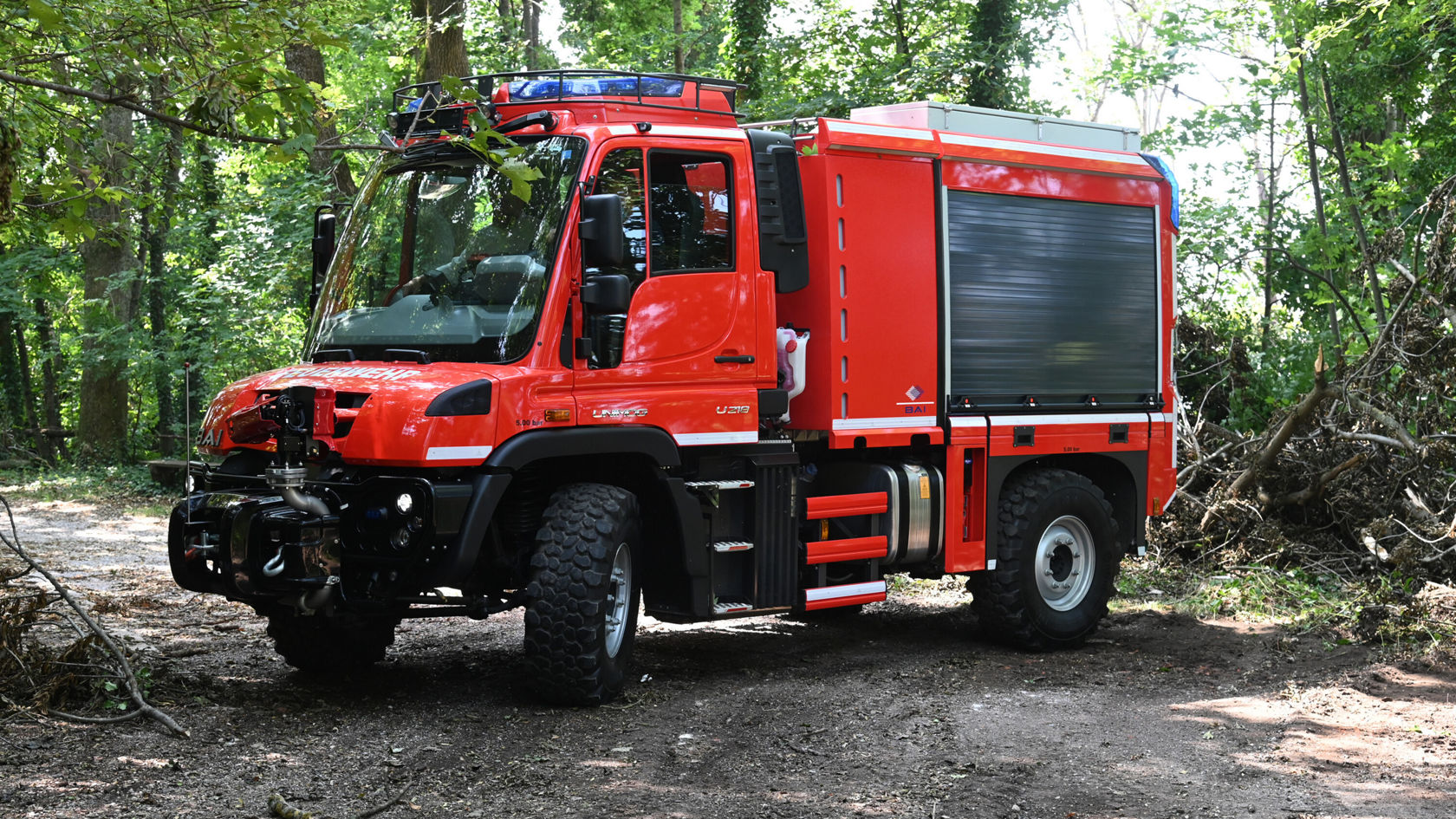Un camion de pompiers Mercedes‑Benz rouge est stationné sur un chemin forestier, équipé d'un gyrophare et d'une plate-forme de chargement fermée, entouré d'arbres et de branchages verts.