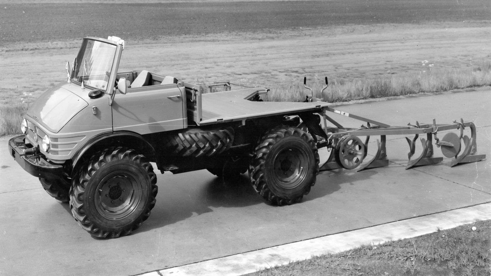 Unimog von Mercedes-Benz mit offener Kabine und breiten Reifen, ausgestattet mit einem Pflug, bereit für landwirtschaftliche Arbeiten auf einem befestigten Weg.