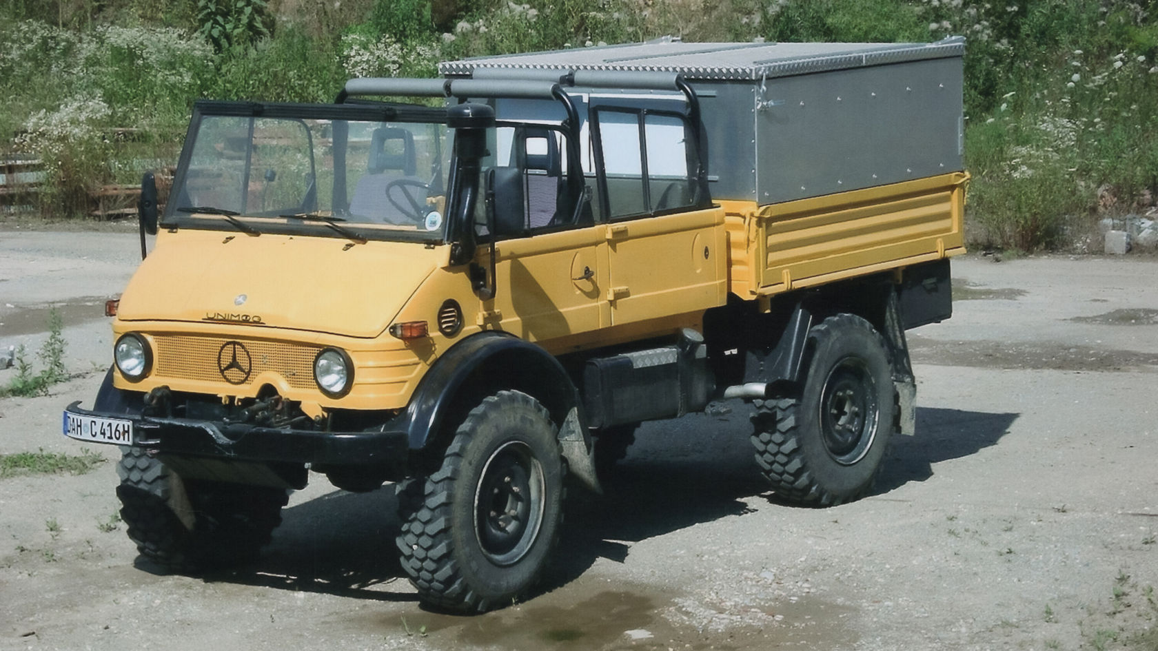 Gelber Unimog von Mercedes-Benz mit offener Kabine und Ladefläche, geparkt auf einer unbefestigten Fläche, optimal für vielseitige Geländearbeiten.