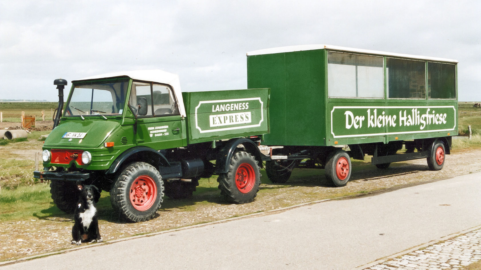 Grüner Mercedes-Benz Unimog mit Anhänger, beschriftet mit „Der kleine Halligfriese“, steht auf einer Straße, daneben sitzt ein schwarzer Hund.