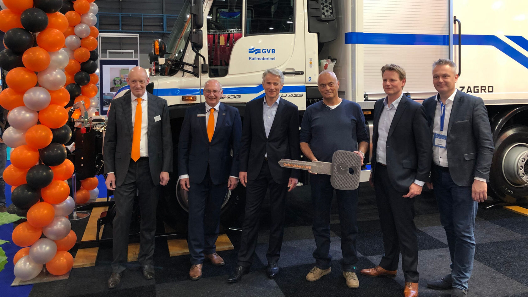 Sechs Männer stehen vor einem Mercedes-Benz Unimog, während einer von ihnen einen großen Schlüssel in der Hand hält. Sie wirken feierlich und lächeln.