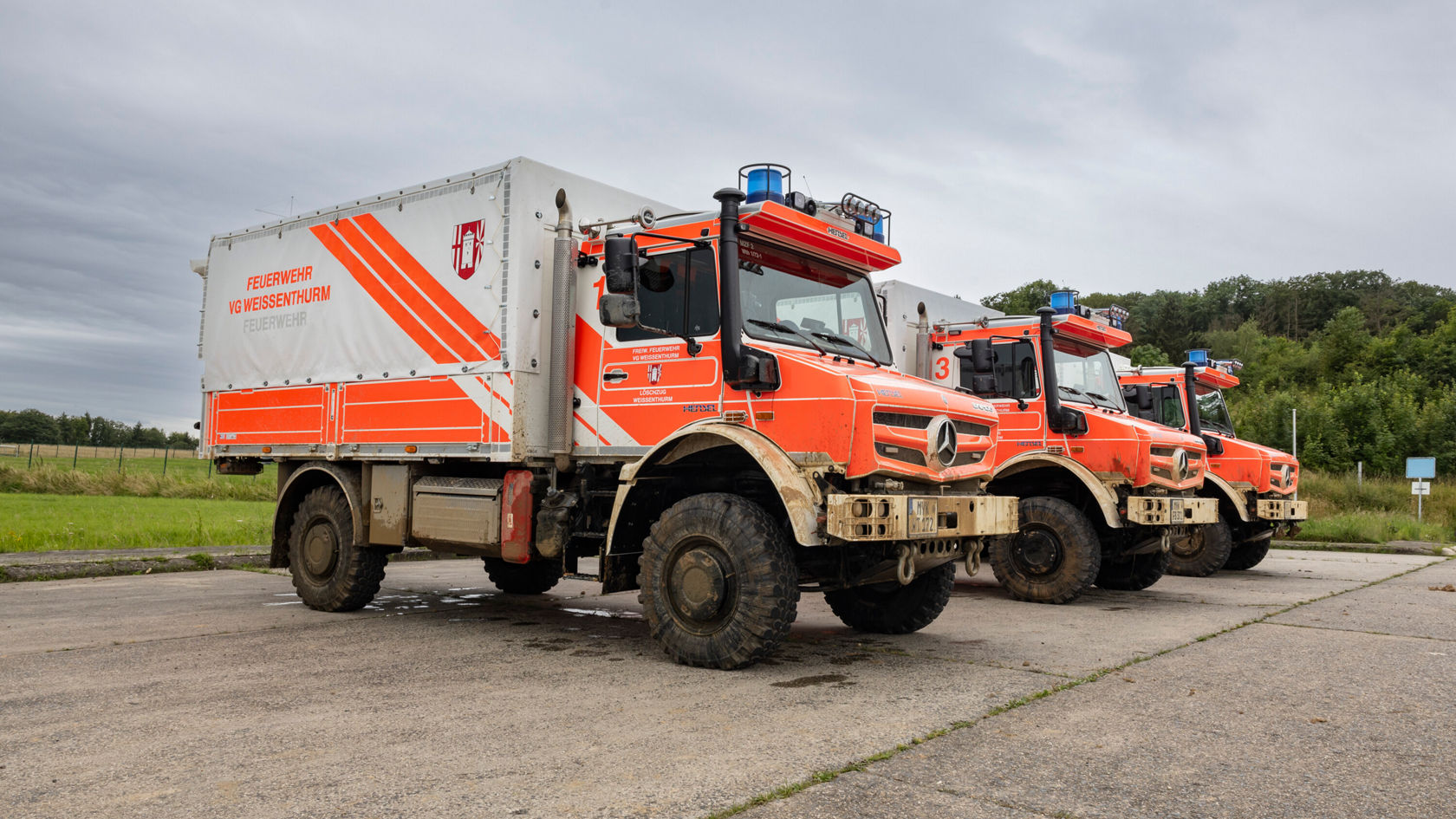 Trois Unimog Mercedes‑Benz orange se trouvent sur une place. Ils sont équipés de gyrophares et disposent d'un plateau de chargement spacieux pour des interventions techniques.