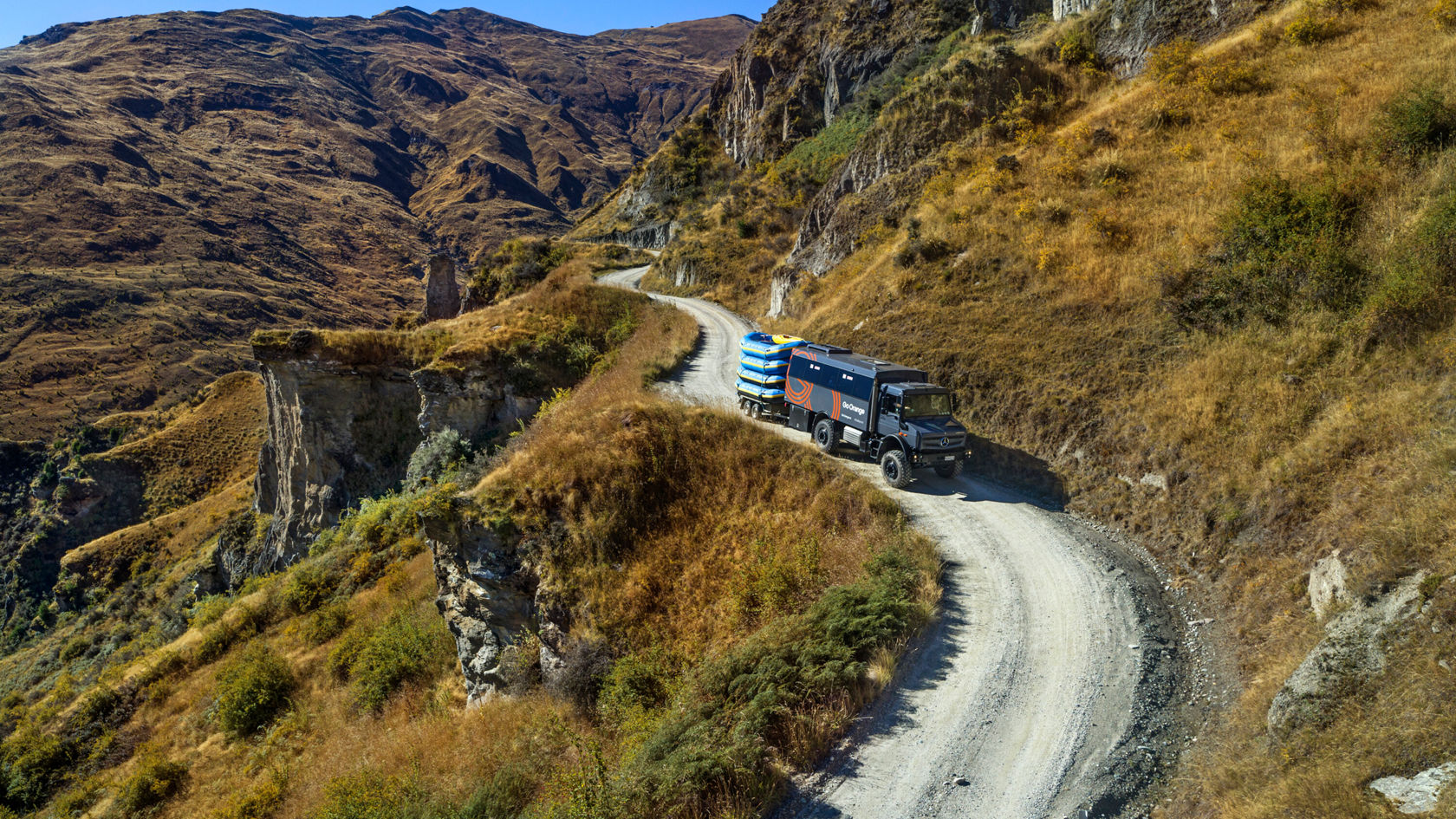 Ein Mercedes-Benz Truck fährt auf einer kurvenreichen Schotterstraße in einer bergigen Landschaft mit trockener Vegetation und felsigen Klippen im Hintergrund.