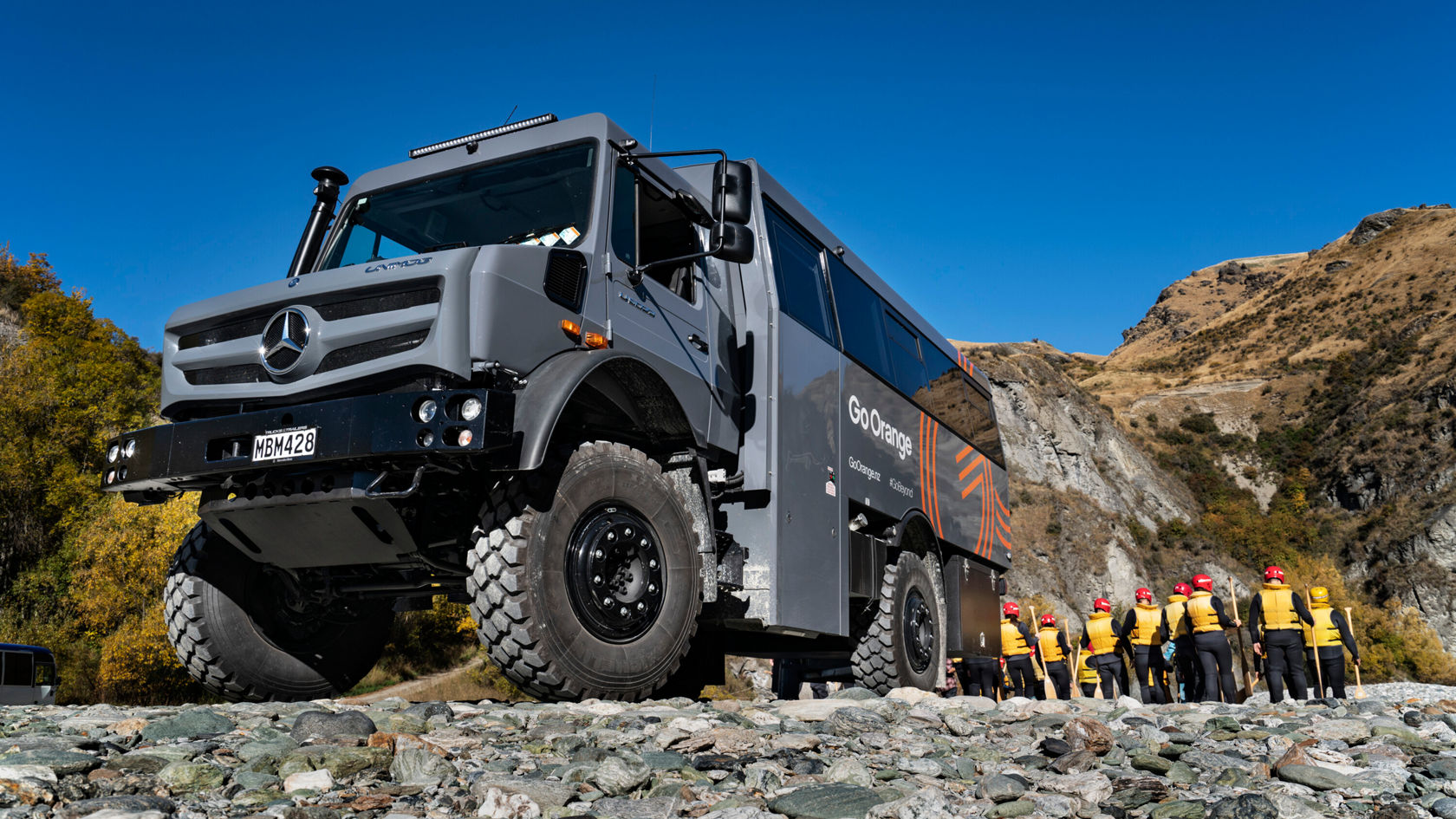 Ein Mercedes-Benz Offroad-Bus steht auf einem Kiesweg. Im Hintergrund stehen eine Gruppe von Personen in gelben Jacken und roten Helmen, die auf den Bus blicken.