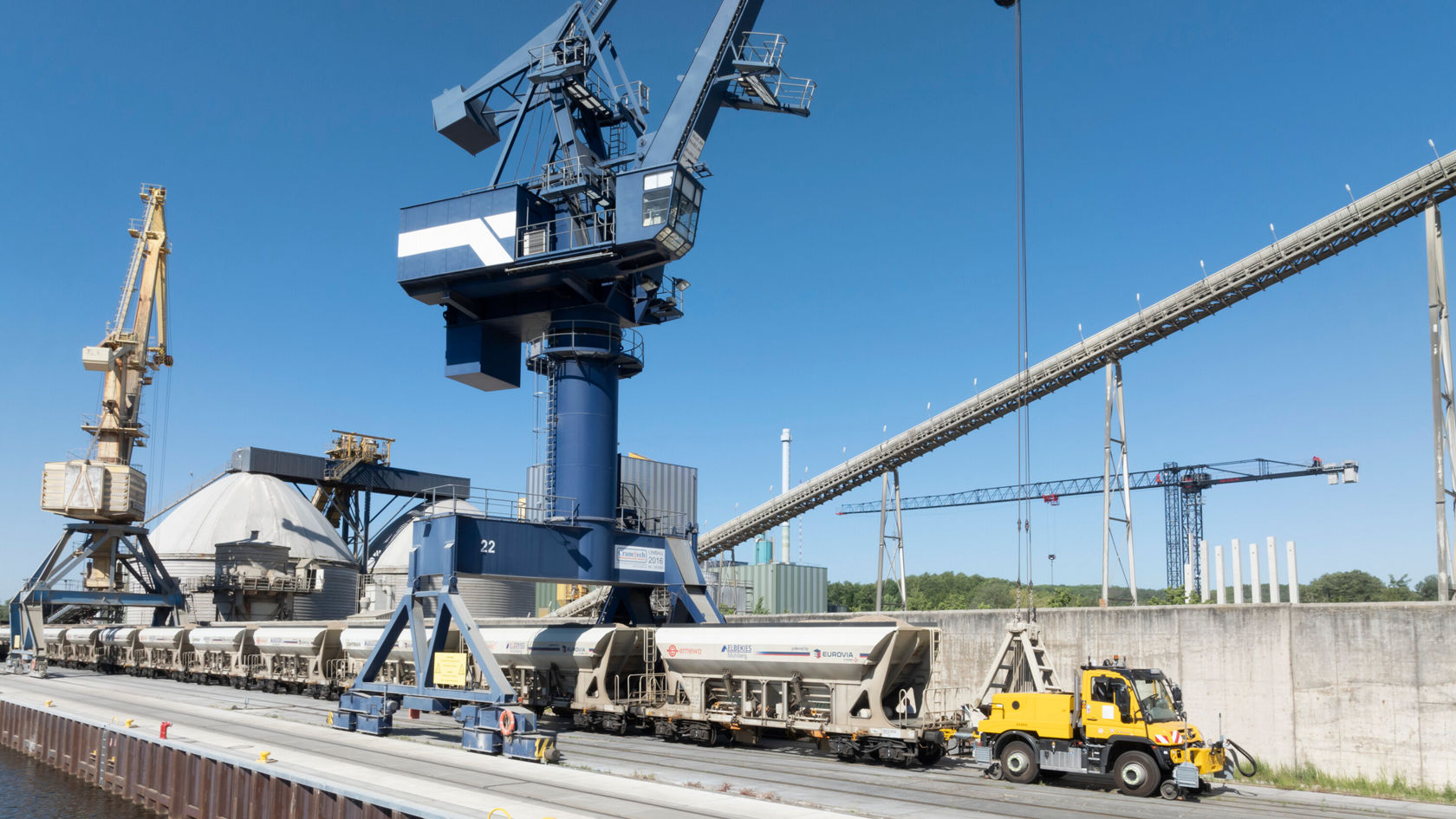 Ein Mercedes-Benz Unimog steht neben einem Güterwagen am Hafen, während ein Kranausleger im Hintergrund Material entlädt. Es ist ein klarer Tag mit blauer Himmel.