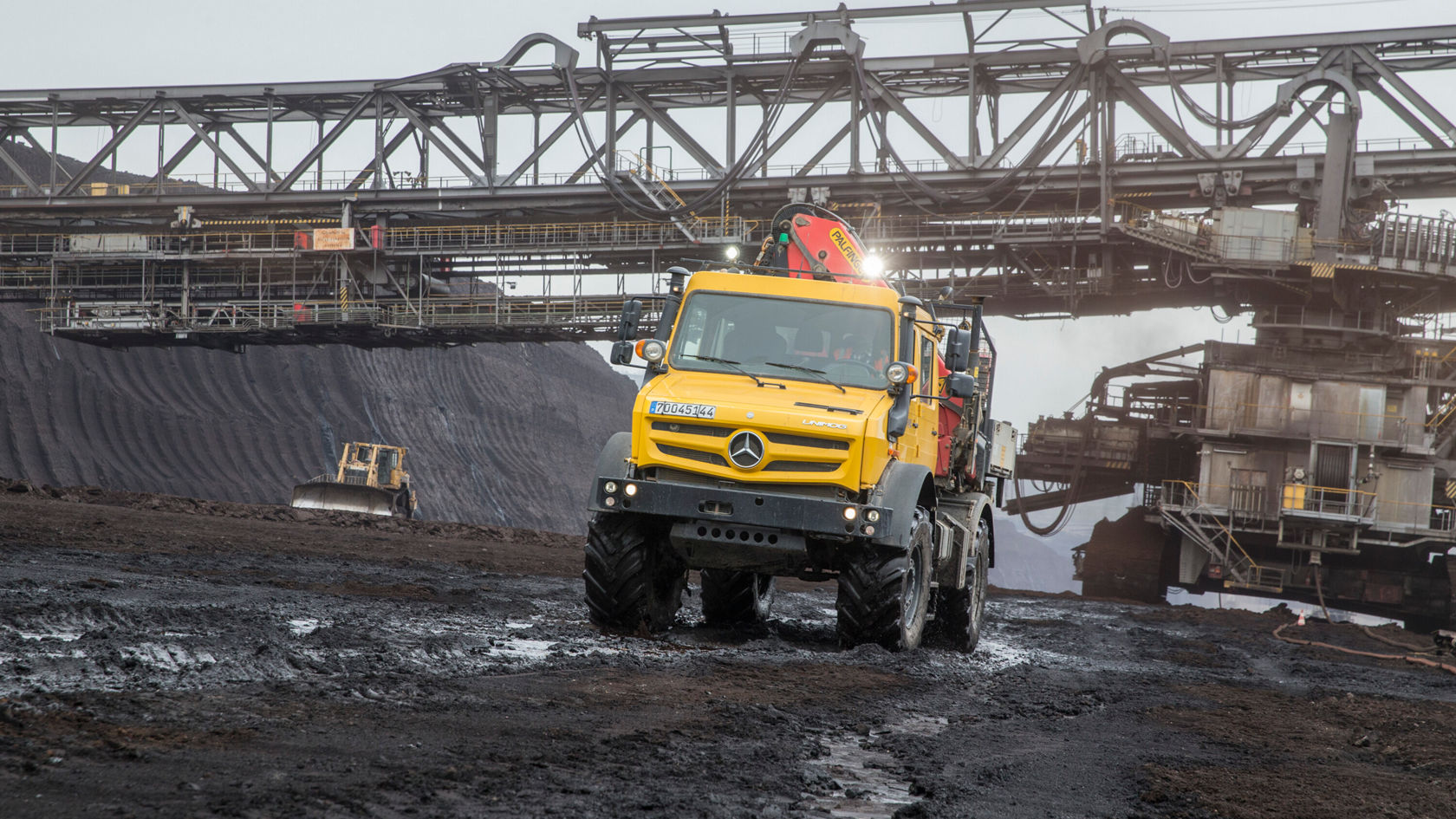 A yellow Mercedes-Benz truck drives across uneven, dirt-covered terrain in an open-cast mine, surrounded by large conveyor systems in the background.