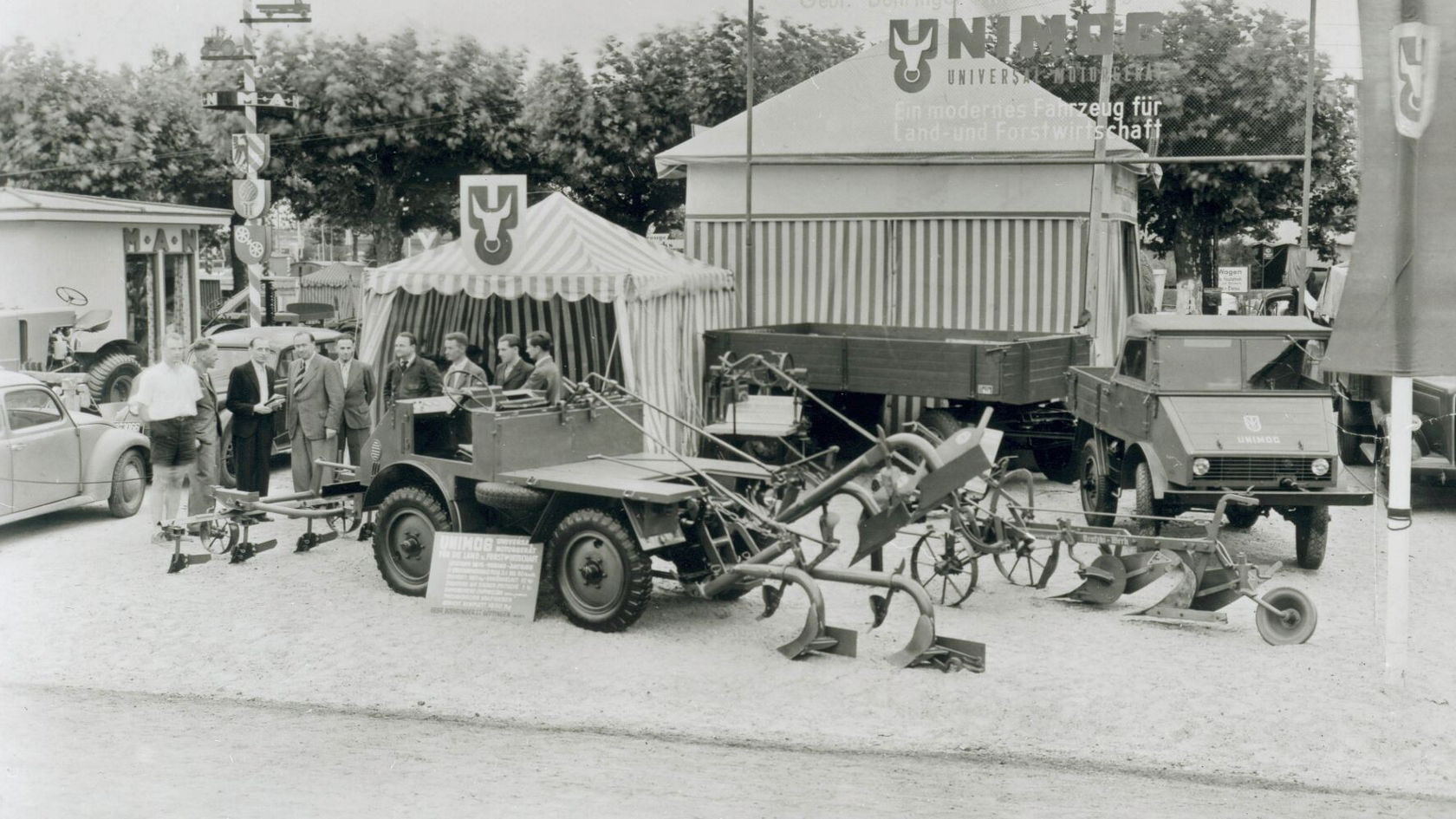 Ein Mercedes-Benz Unimog steht auf einer Messe, umgeben von landwirtschaftlichen Geräten und mehreren Personen, die das Fahrzeug und die Ausrüstung besprechen.
