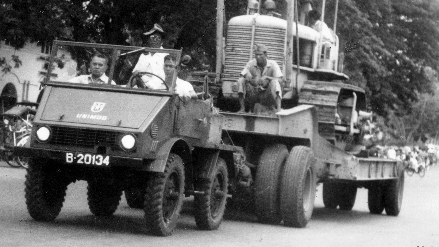 Ein Mercedes-Benz Unimog zieht einen Anhänger mit einem Traktor, während mehrere Personen im Fahrzeug sitzen. Es handelt sich um eine historische Aufnahme mit einem ländlichen Hintergrund.