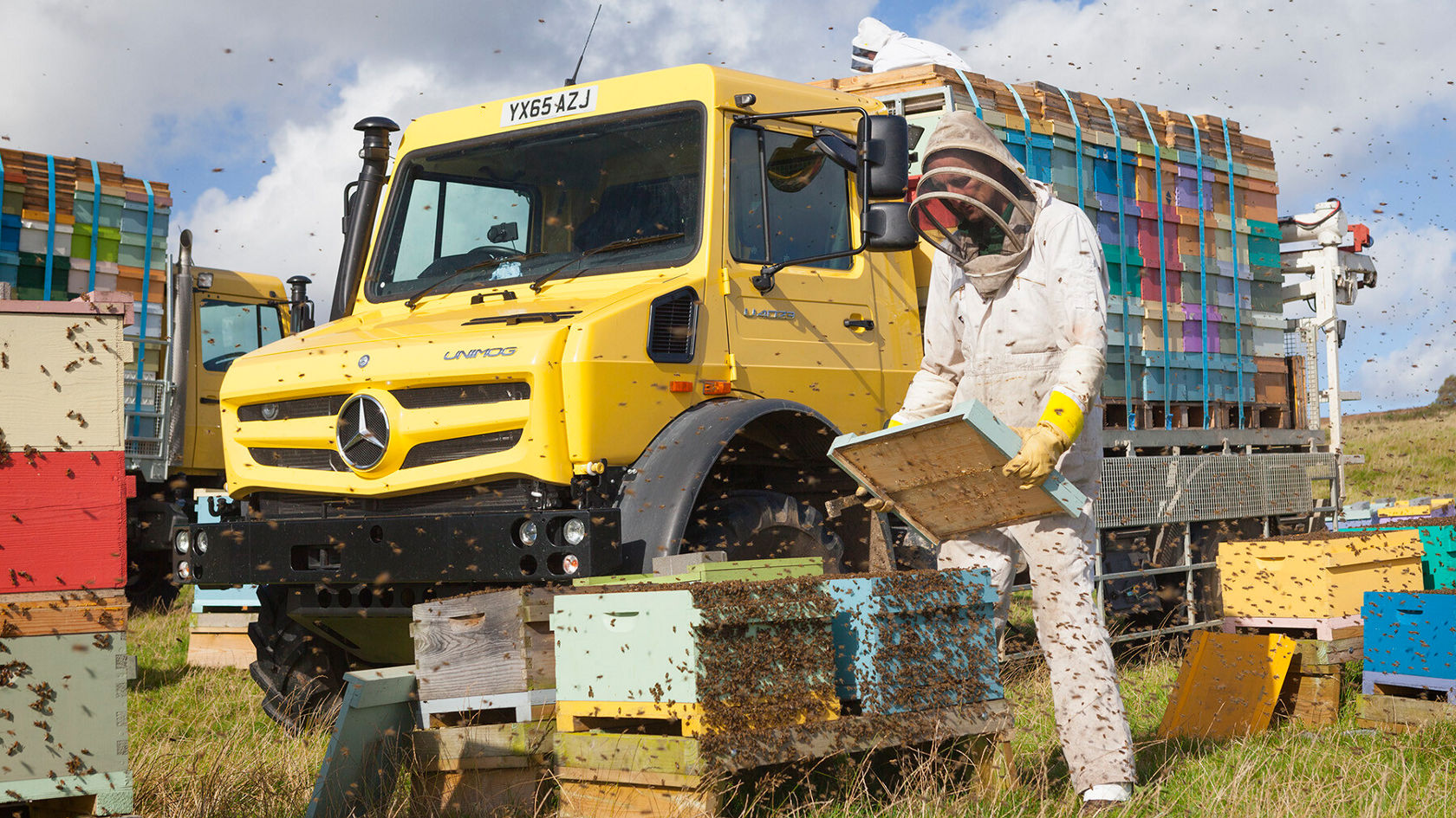 Ein gelber Unimog von Mercedes-Benz steht neben einem Imker in Schutzkleidung, der Bienenstöcke mit einem Rahmen inspiziert. Bienen fliegen um ihn herum.