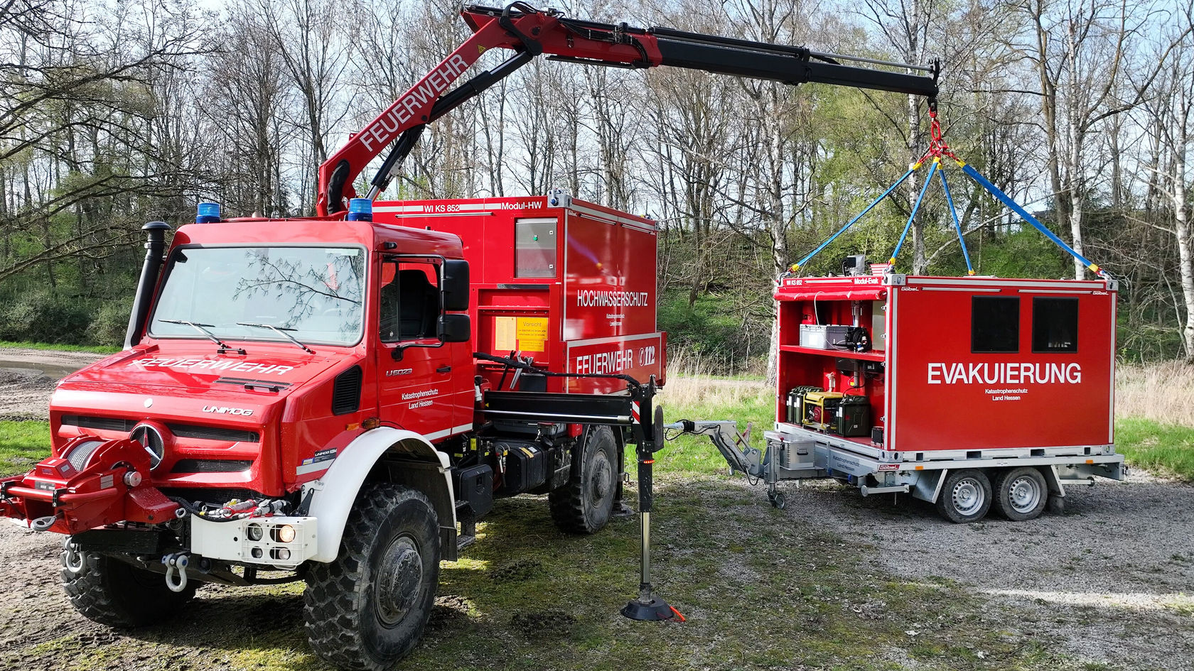 Un camion de pompiers Mercedes‑Benz rouge équipé d'une grue soulève une remorque portant l'inscription « ÉVACUATION ». La scène montre une situation d'intervention en plein air.