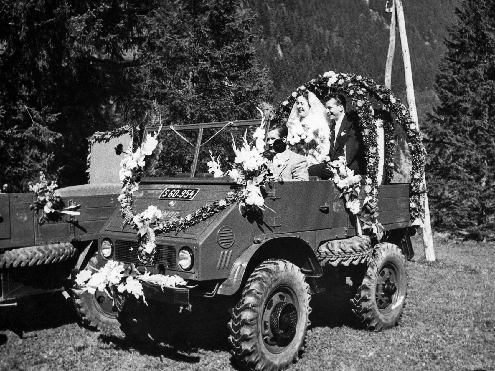 Huwelijksfeest in een met bloemen versierde Mercedes-Benz Unimog, met een bruidspaar en andere gasten, omgeven door bomen op een weide.