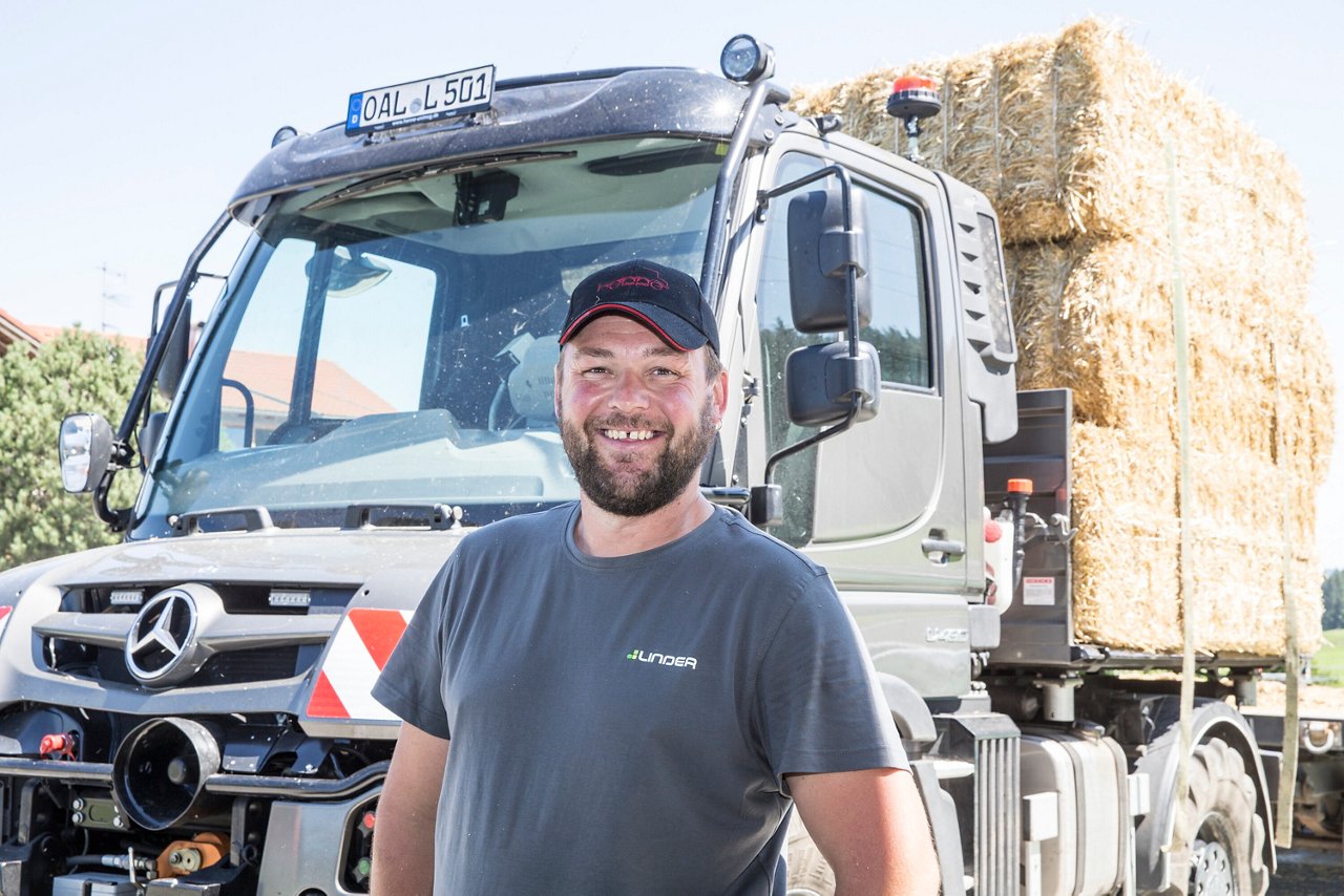 Un homme satisfait se tient devant un camion Mercedes‑Benz chargé de balles de foin. La scène transmet une impression de travail rural et d'activité de transport agricole.