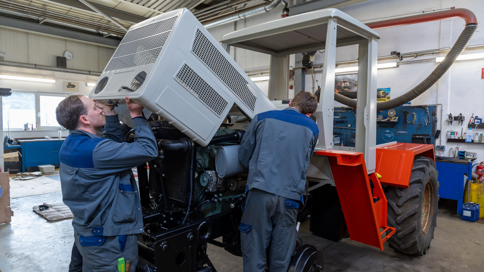 Zwei Mechaniker arbeiten in einer Werkstatt an einem Mercedes-Benz Unimog, während sie eine Abdeckung auf den Motor setzen.