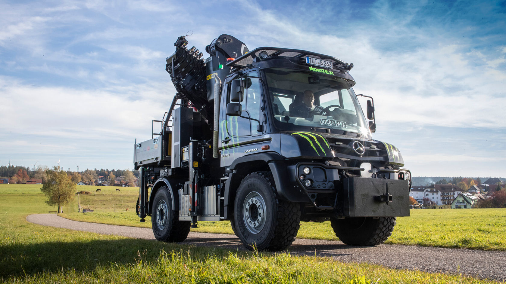 Un Unimog Mercedes‑Benz doté d'une superstructure de grue est arrêté sur un chemin de campagne, entouré de prairies et d'arbres, sous un ciel dégagé.
