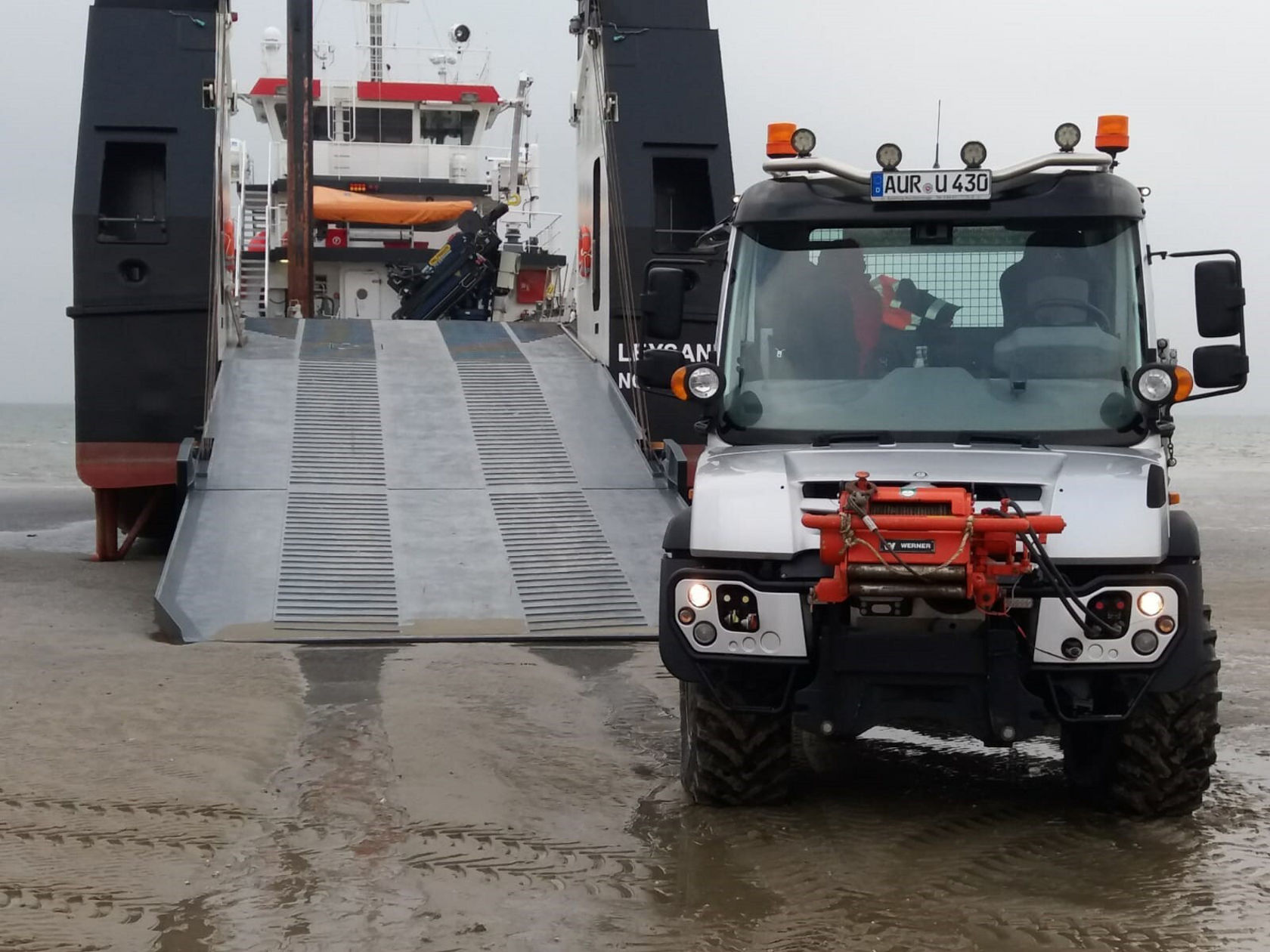 Un Unimog Mercedes‑Benz se trouve sur le sable, tandis qu'un ferry accoste. La rampe du ferry est abaissée pour recevoir le véhicule.