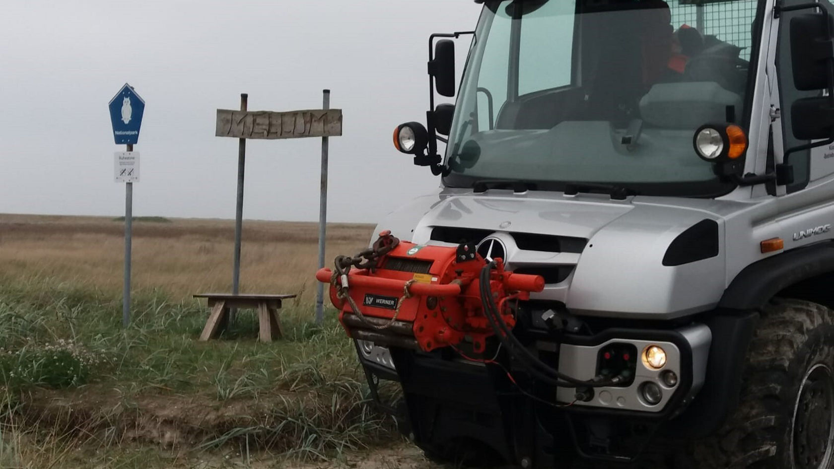 Un Unimog Mercedes‑Benz est stationné sur un chemin de terre à côté d'un panneau en bois portant l'inscription « MELLUM ». Un panneau d'arrêt est également visible.