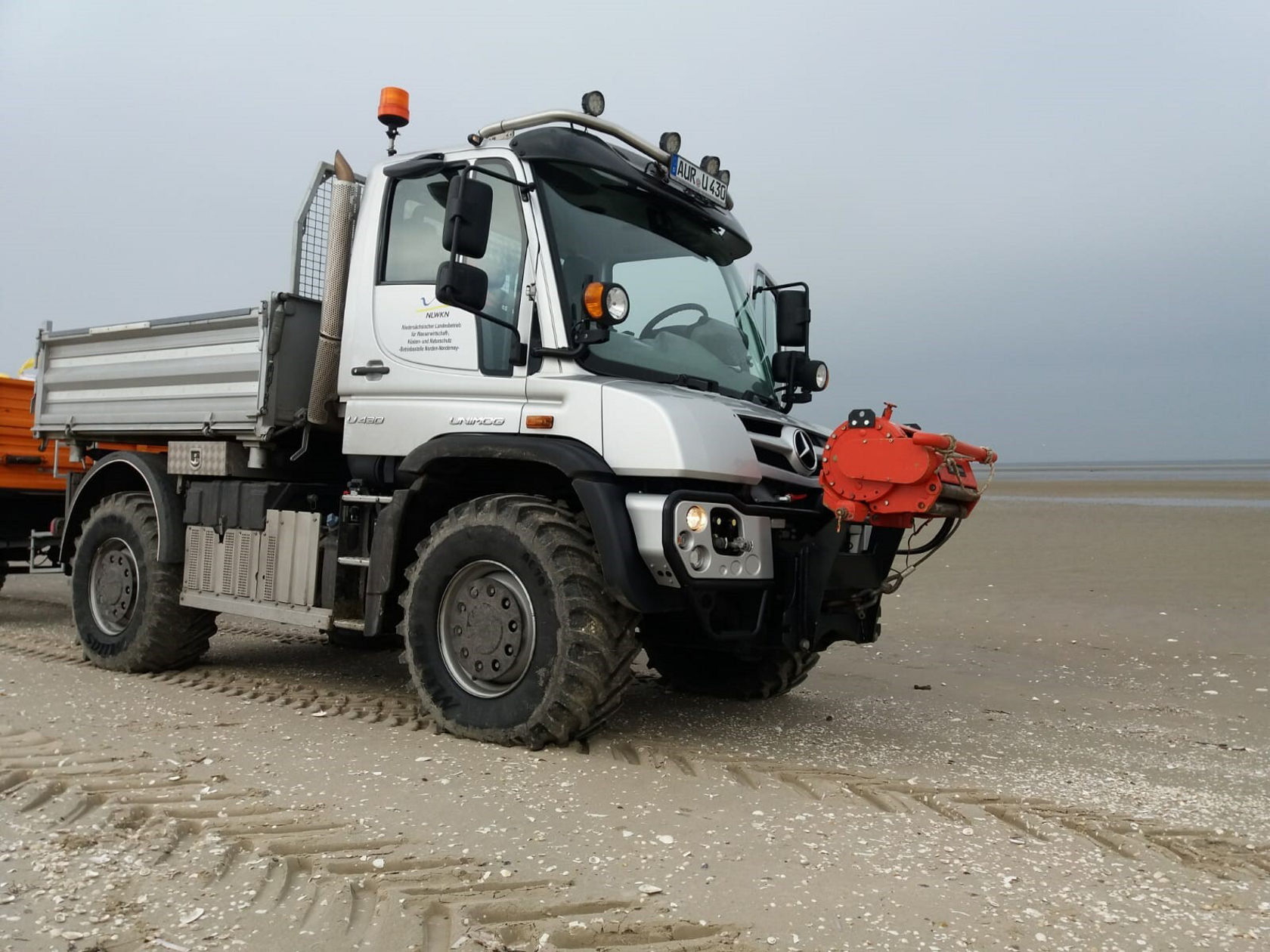 Un Unimog Mercedes‑Benz est stationné sur un terrain sablonneux, équipé d'un outil rapporté rouge à l'avant et d'une zone de chargement ouverte à l'arrière, prêt pour des interventions tout‑terrain.