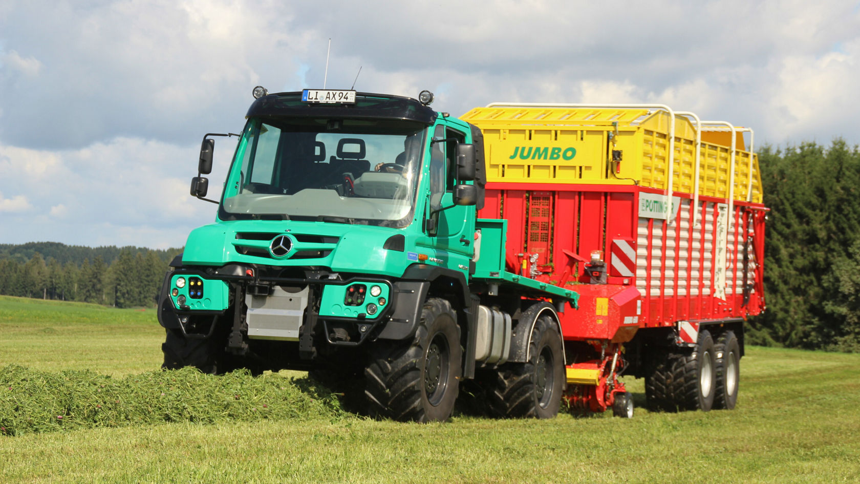Un tractor Mercedes-Benz con un remolque Jumbo rojo circula por un campo, rodeado de un paisaje verde. El tractor tiene neumáticos grandes y un color turquesa llamativo.