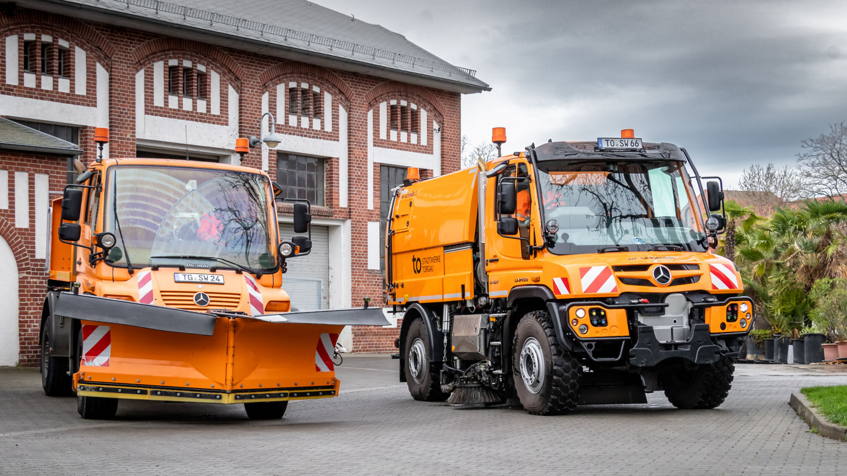 Zwei orangefarbene Mercedes-Benz Unimog-Fahrzeuge stehen in einer städtischen Umgebung. Das linke Fahrzeug ist mit einer Schneeschaufel ausgestattet, während das rechte Fahrzeug für Reinigungsarbeiten bereit ist.