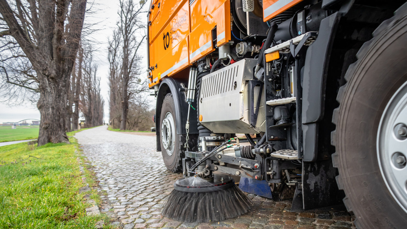 Ein Unimog von Mercedes-Benz reinigt einen gepflasterten Weg mit einer Bürste. Die Szene zeigt das Fahrzeug in Aktion, umgeben von Bäumen und Gras.