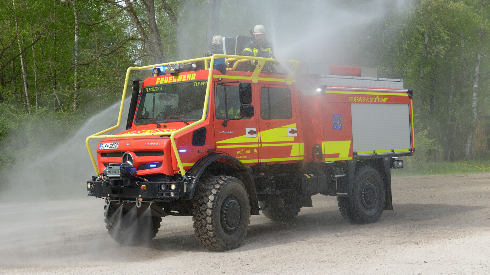 Un Unimog Mercedes‑Benz rouge projette de l'eau, tandis qu'un pompier se tient sur le toit.