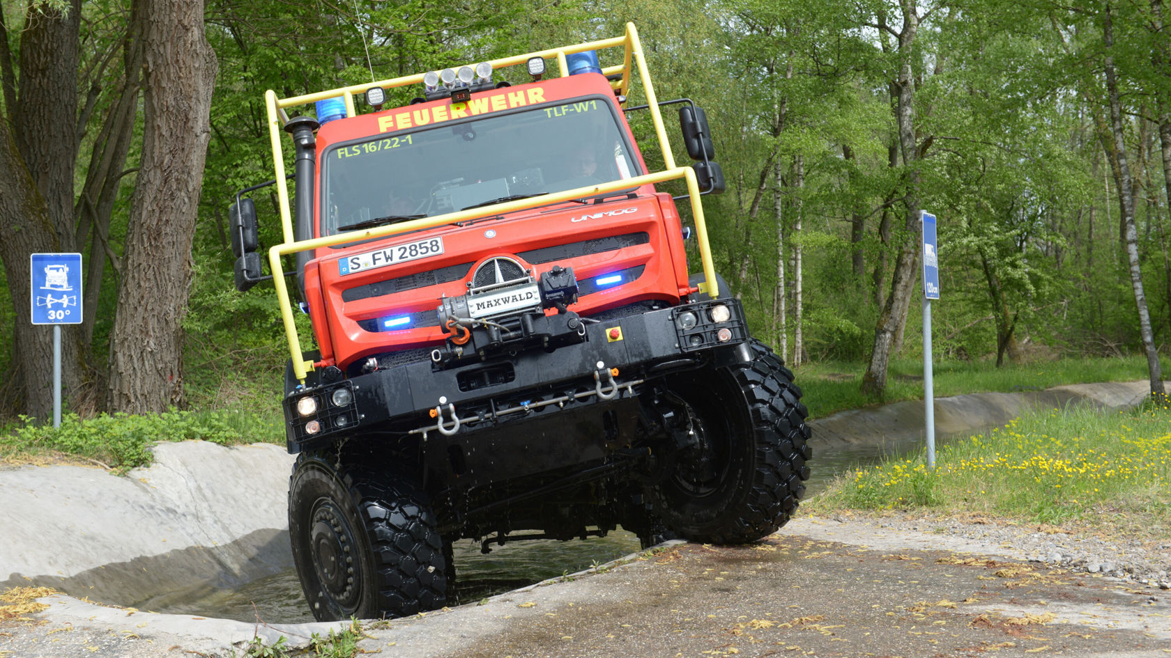 Un Unimog Mercedes‑Benz rouge se trouve sur un terrain accidenté et montre ses capacités tout-terrain, tandis que le gyrophare est allumé.