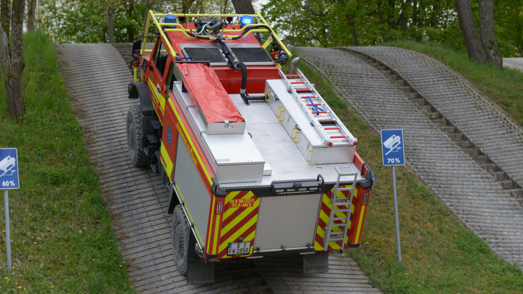 Un Unimog Mercedes‑Benz rouge monte une rampe raide, équipé d'une superstructure d'intervention. Des panneaux sur le côté indiquent des pentes de 60 % et 70 %.