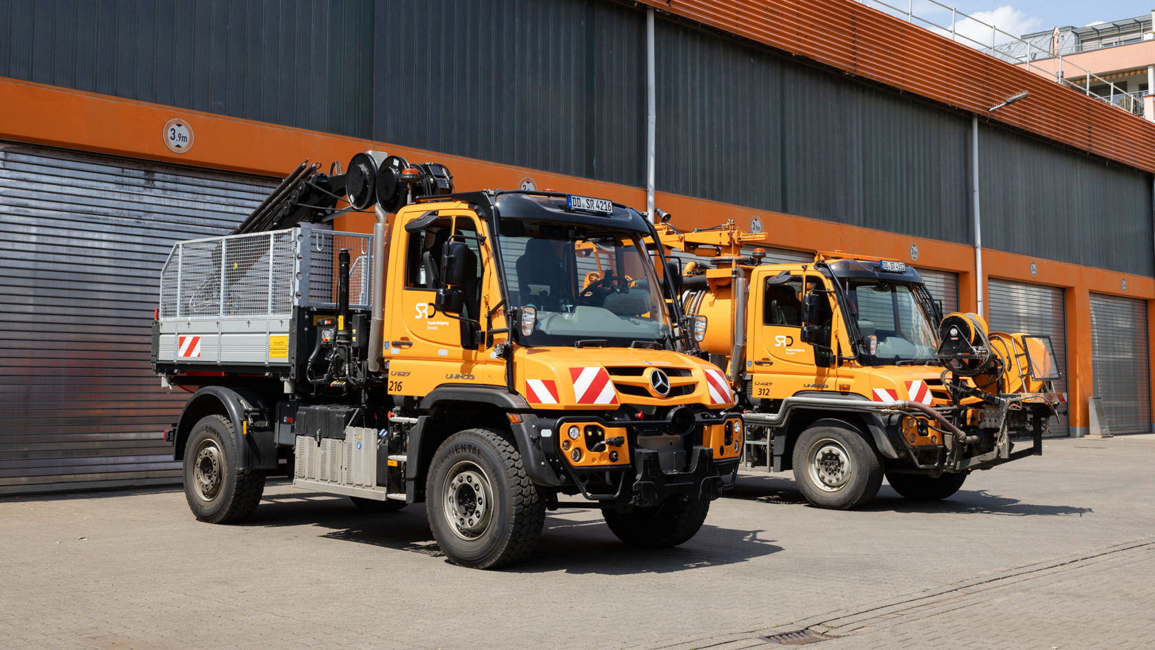 Two yellow Mercedes-Benz Unimog vehicles are parked in front of a warehouse. The vehicles are equipped with tools and loads for construction or maintenance work.