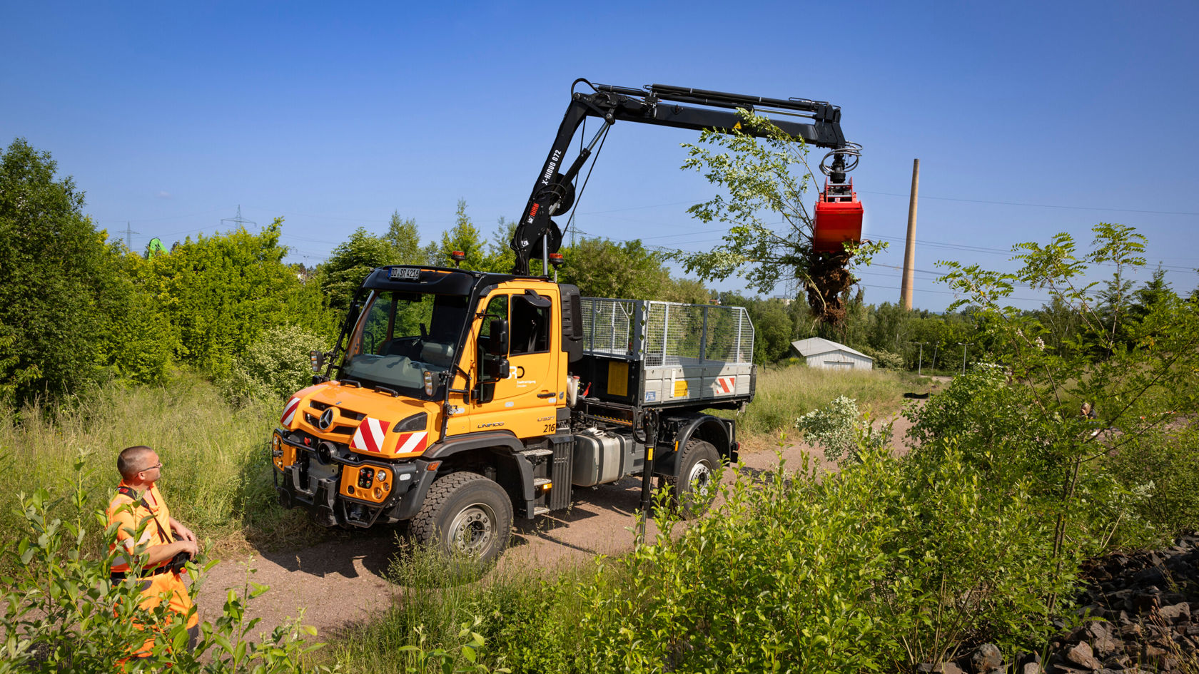 A Mercedes-Benz Unimog with a crane is lifting a tree trunk while a worker stands next to it, surrounded by greenery and trees on an unpaved road.