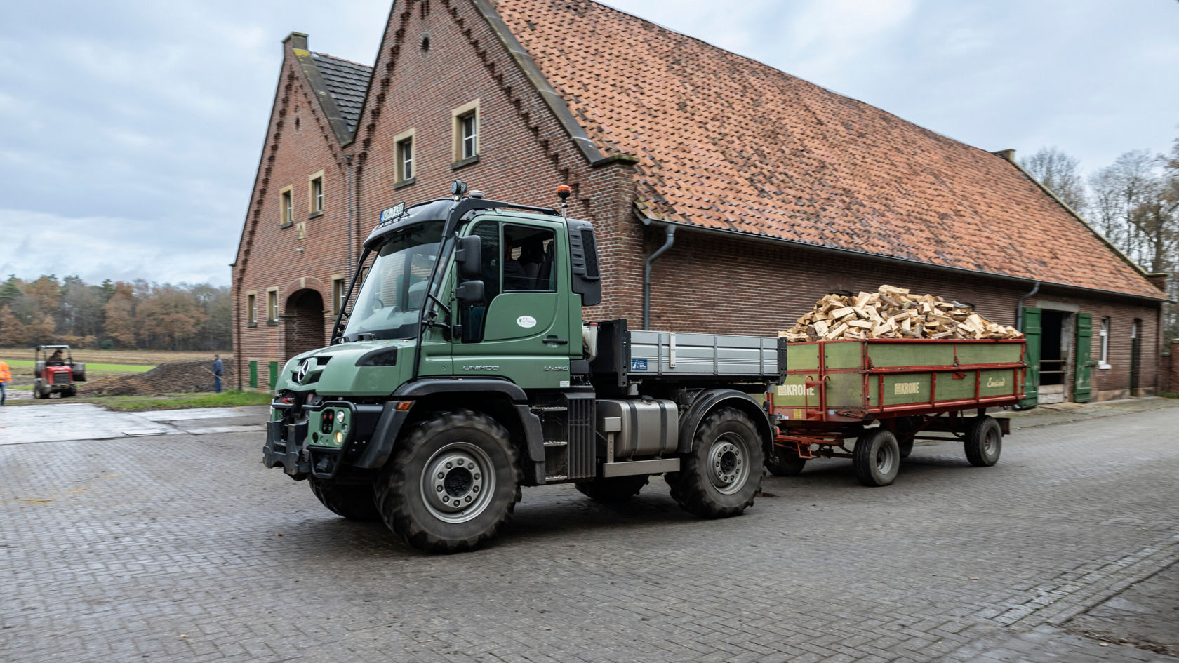 A green Unimog with a trailer, loaded with wood, stands in front of an old farmhouse.