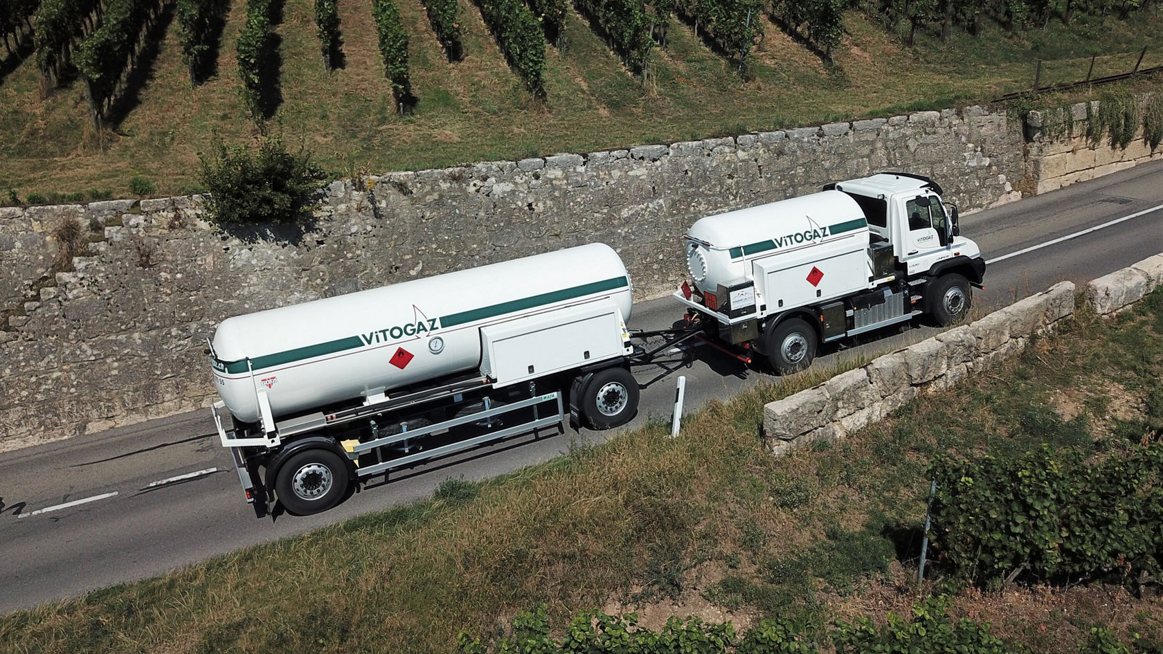 Ein Mercedes-Benz-Lkw transportiert einen großen weißen Tank für Vitogaz auf einer Straße, umgeben von Weinbergen und einer Steinmauer.