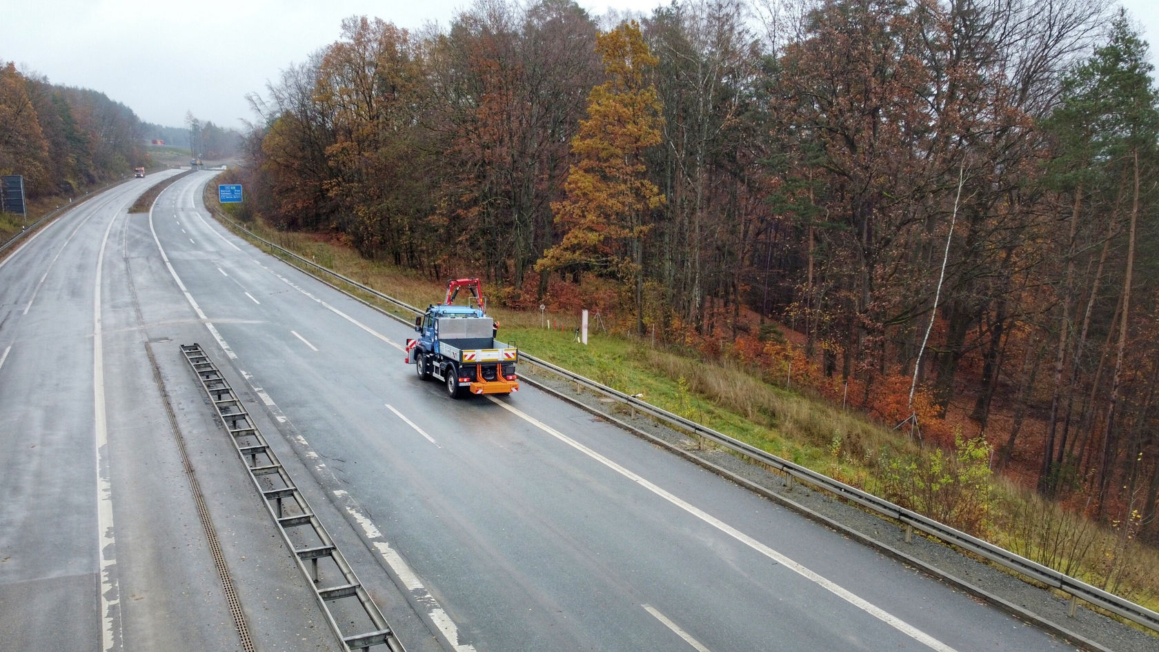 Un Unimog Mercedes‑Benz circule sur une route sous la pluie et est équipé d'un gyrophare rouge afin d'assurer les mesures de sécurité lors de travaux routiers.