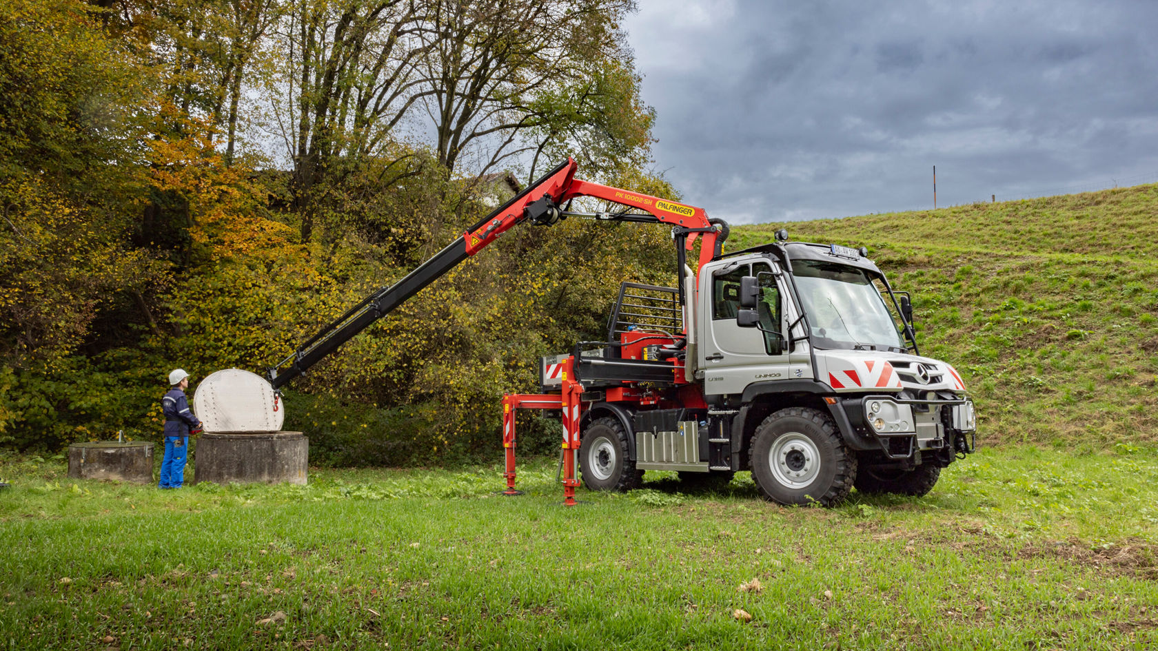 Un Unimog Mercedes‑Benz muni d'un bras de grue soulève une grande plaque ronde sur un terrain, avec des arbres en arrière-plan. Un ouvrier en vêtements bleus surveille l'opération.