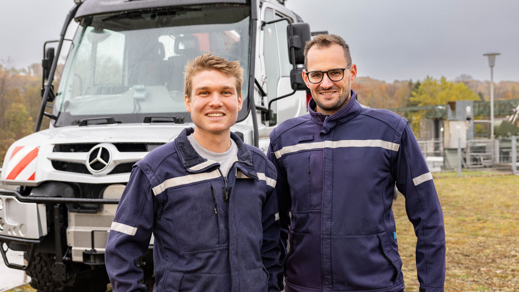 Deux mécaniciens en tenues de travail bleues se tiennent, souriants, devant un véhicule Mercedes‑Benz. L'environnement semble industriel, avec des arbres en arrière-plan.