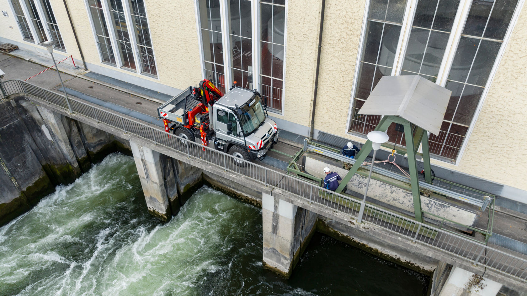Un véhicule Mercedes‑Benz se trouve sur une passerelle à côté d'une centrale hydroélectrique, tandis que de l'eau coule sous la passerelle. La passerelle est fonctionnelle et bien éclairée.