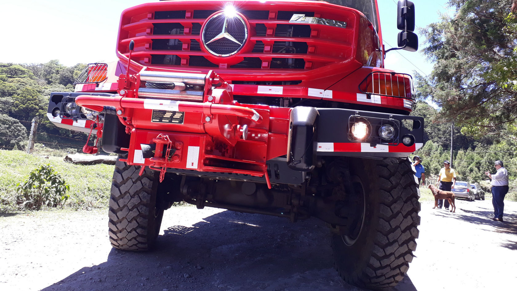 Un Unimog Mercedes-Benz rouge est stationné sur une route non revêtue. Au premier plan, on voit de gros pneus et une face avant frappante, tandis que des personnes se tiennent en arrière-plan.